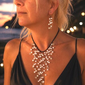 May include: A necklace and matching earring set featuring white pearls and small metallic floral accents suspended on thin black threads. The necklace has a cascading, multi-strand design that drapes across the chest, while the earring is a drop style.