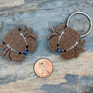 May include: Two brown metal crab keychains with blue accents. The keychains are attached to a silver key ring.
