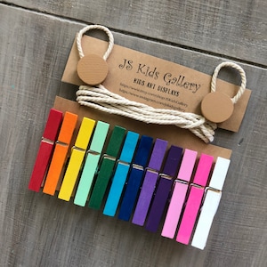 May include: A set of 15 colorful wooden clothespins, arranged in a rainbow pattern, with a white rope and two brown wooden circles. The text "JS Kids Gallery" and "Kids Art Displays" is printed on a brown paper background. The website addresses "https://www.etsy.com/shop/JSKidsGallery" and "https://www.instagram.com/jskidsgallery" are also printed on the paper.