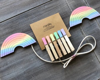 May include: A set of rainbow-colored wooden clothespins with a string attached to two rainbow-shaped wooden cutouts. The string is tied in a bow. The clothespins are arranged in a row on a brown card with the text "JS Kids Gallery" printed on it.