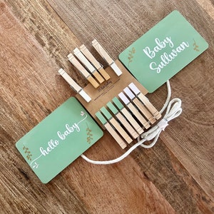 May include: A set of wooden clothespins with a green and white string, used to hang two green signs with the text "hello baby" and "Baby Sullivan".