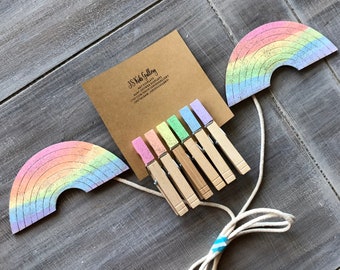 May include: A set of rainbow-colored wooden clothespins with a string and two rainbow-shaped wooden decorations. The clothespins are decorated with pastel colors and glitter. The string is white with a blue and white stripe. The rainbow decorations are made of wood and have a glitter finish.