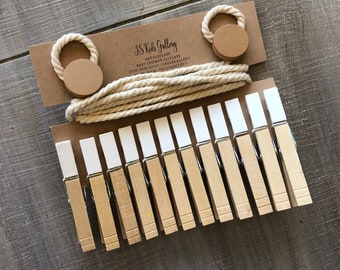May include: A set of 15 white wooden clothespins with a natural wood finish. The clothespins are attached to a length of natural twine with two brown wooden circles at the ends. The twine is wrapped around a brown card with the text "JS Kids Gallery Art Displays Baby Shower, Nursery, Etsy.com/shop/JSKidsGallery Instagram: JSKidsGallery".