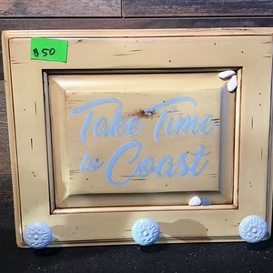 May include: A light yellow wooden cabinet door with a white sign that says "Take Time to Coast". The door has three blue knobs and white decorative accents.