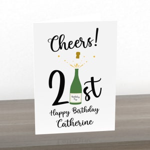 Puede incluir: Una tarjeta blanca con texto negro que dice "Cheers! 21st Happy Birthday Catherine". Hay una botella de champ&aacute;n verde con una tapa dorada que se abre con chispas doradas.