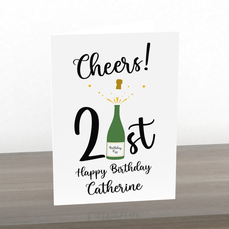 Puede incluir: Una tarjeta blanca con texto negro que dice "Cheers! 21st Happy Birthday Catherine". Hay una botella de champ&aacute;n verde con una tapa dorada que se abre con chispas doradas.