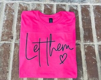 Könnte beinhalten: Pinkes T-Shirt mit dem Text "Let them" in schwarzer Schrift mit einem Herz am Ende.