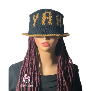 Pode incluir: Um chapéu de balde de crochê preto com um "YAH" bordado em dourado na frente. O chapéu é usado por uma pessoa com longos dreadlocks bordô.
