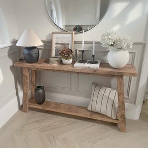 May include: A wooden console table with a lower shelf, displaying a lamp, framed art, candles, and a vase of white flowers. A striped cushion rests on the lower shelf. A round mirror is in the background.