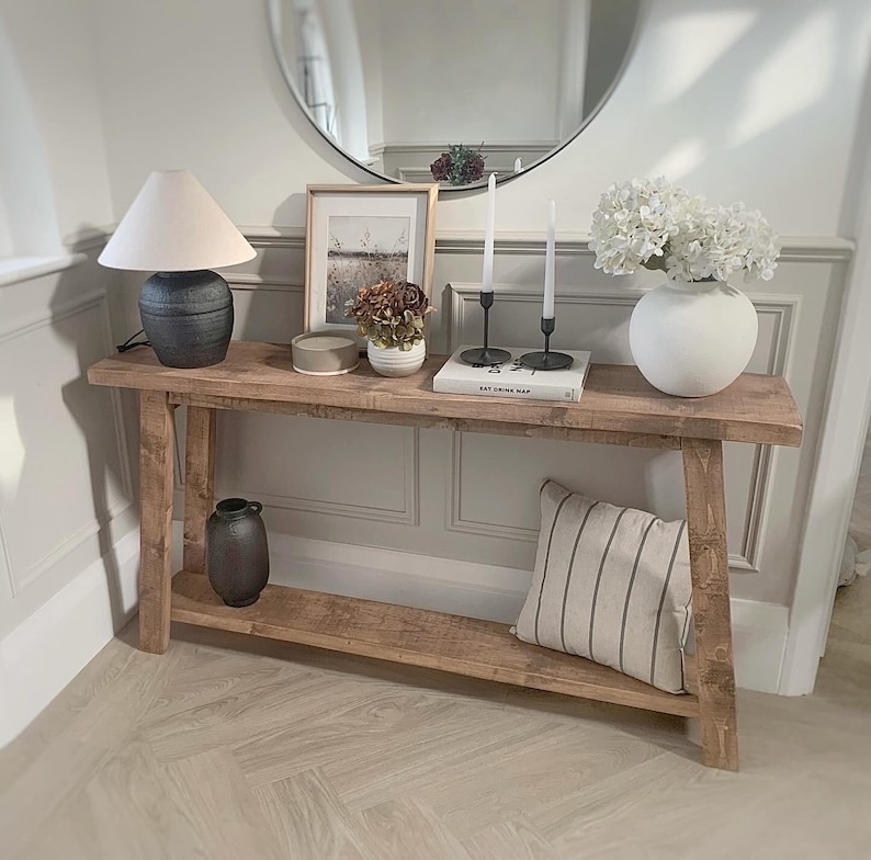 K&ouml;nnte beinhalten: Ein rustikaler Holztisch mit unterem Regal, auf dem eine Lampe, gerahmte Kunst, Kerzen und eine Vase mit wei&szlig;en Blumen stehen. Ein gestreiftes Kissen liegt auf dem unteren Regal. Im Hintergrund ist ein runder Spiegel zu sehen.