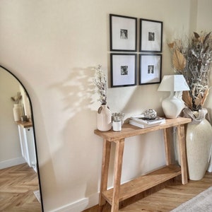 May include: A light wood console table with a lower shelf, featuring a white vase with dried flowers, a candle, and a stack of books. The table is against a white wall with black framed photos.
