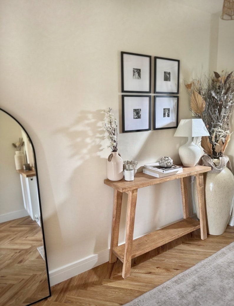 May include: A light wood console table with a lower shelf, featuring a white vase with dried flowers, a candle, and a stack of books. The table is against a white wall with black framed photos.