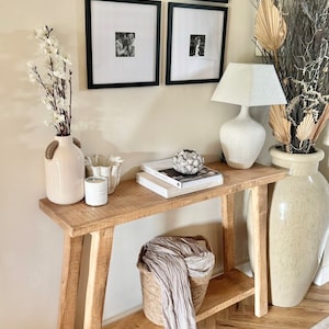 May include: A wooden console table with a shelf, featuring a white lamp with a white shade, a white vase with a floral arrangement, a white bowl, a candle, and a stack of books. The table is set against a white wall with four black framed pictures.