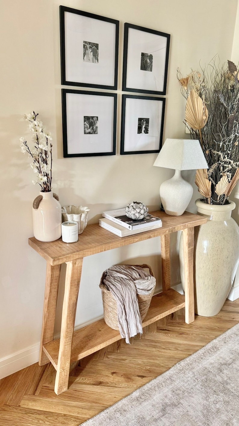 May include: A wooden console table with a shelf, featuring a white lamp with a white shade, a white vase with a floral arrangement, a white bowl, a candle, and a stack of books. The table is set against a white wall with four black framed pictures.