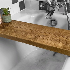 May include: A wooden bath tray with a natural finish, placed across a white bathtub. The tray has two small potted succulents and a bottle of liquid on it.