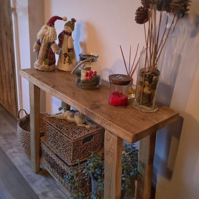 May include: A rustic wooden console table with two shelves. The table is decorated with a snowman and Santa Claus figurine, a glass jar with pine cones and red berries, a glass jar with stones, and a candle in a glass jar.