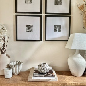 May include: Four black framed photographs of a couple in black and white hang on a white wall above a wooden console table. A white ceramic lamp with a white lampshade sits on the table. A white ceramic artichoke sits on a stack of books on the table.