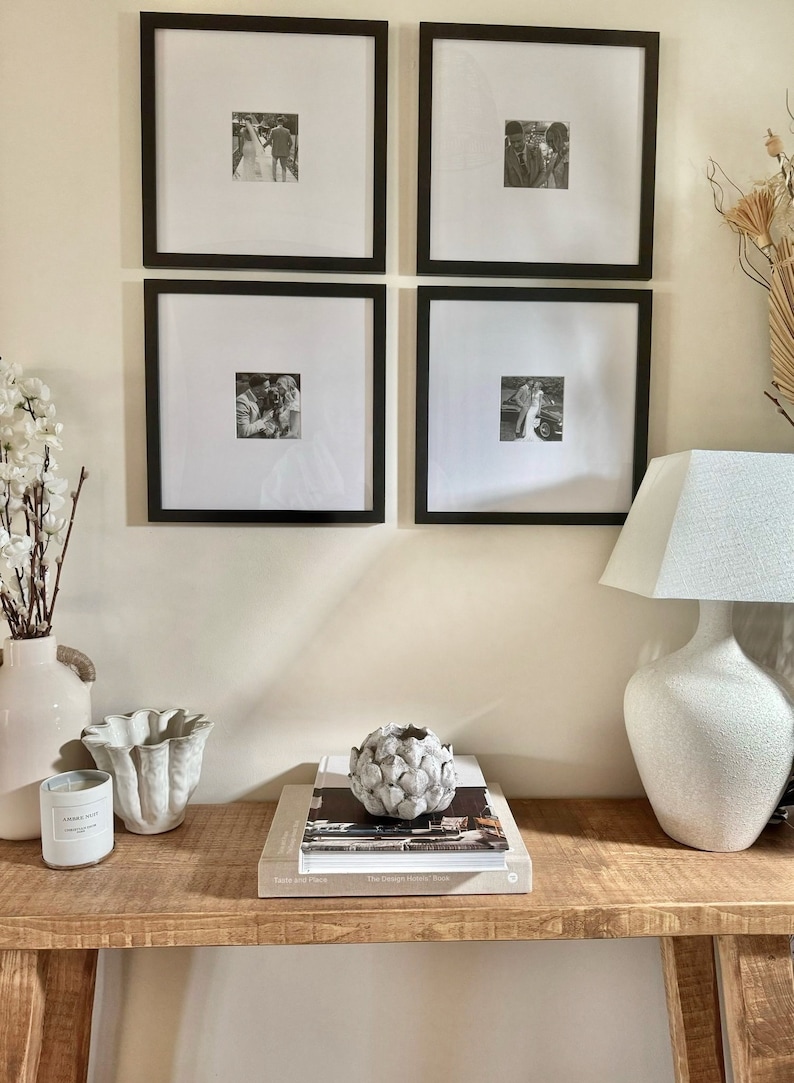 May include: Four black framed photographs of a couple in black and white hang on a white wall above a wooden console table. A white ceramic lamp with a white lampshade sits on the table. A white ceramic artichoke sits on a stack of books on the table.