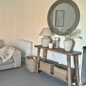 May include: A rustic wooden console table with a white lamp, candles, and a vase of white flowers. The table has two shelves and is decorated with woven baskets.