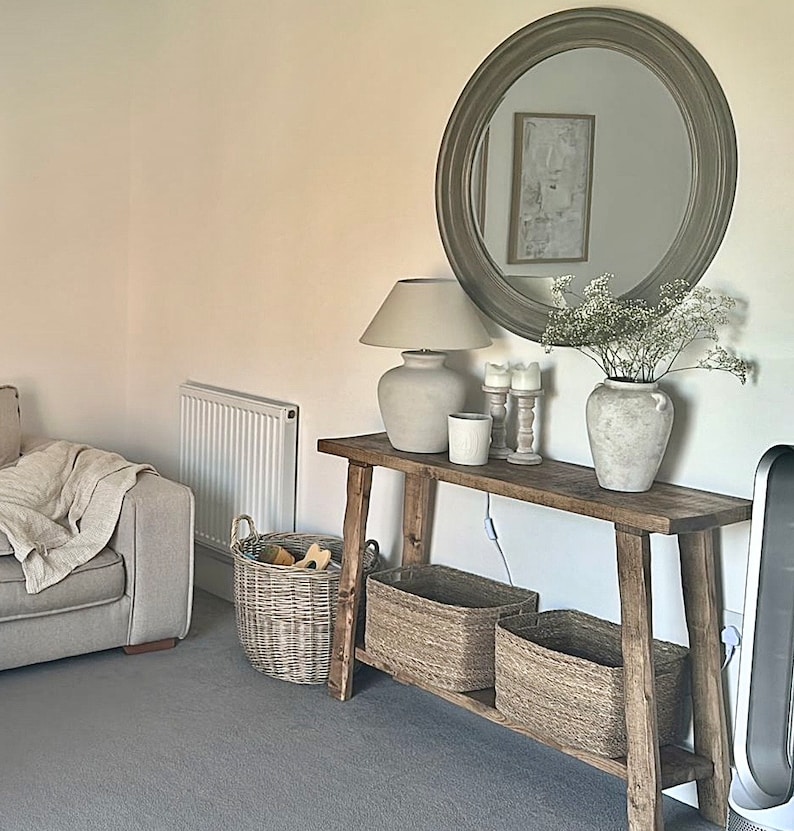 May include: A rustic wooden console table with a white lamp, candles, and a vase of white flowers. The table has two shelves and is decorated with woven baskets.