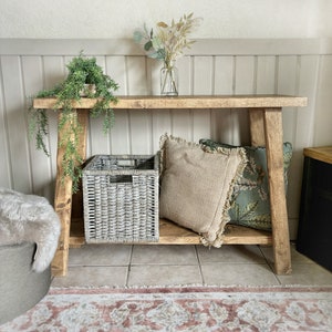 May include: A rustic wooden console table with a grey wicker storage basket, two throw pillows, and a vase of dried flowers. The table has a natural wood finish and is supported by two A-frame legs.