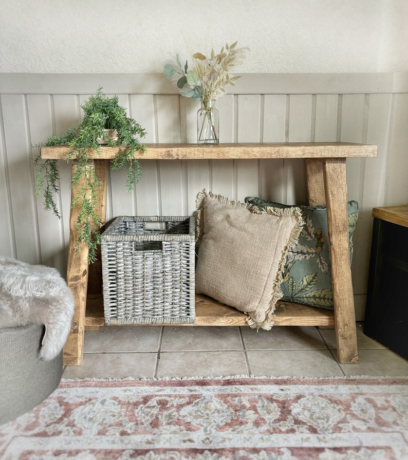 May include: A rustic wooden console table with a grey wicker storage basket, two throw pillows, and a vase of dried flowers. The table has a natural wood finish and is supported by two A-frame legs.