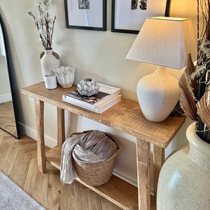 May include: A wooden console table with a shelf, featuring a white ceramic lamp with a linen shade, a white ceramic vase with white flowers, a white ceramic bowl, and a stack of books. The table is in a room with a white wall and a wood floor.