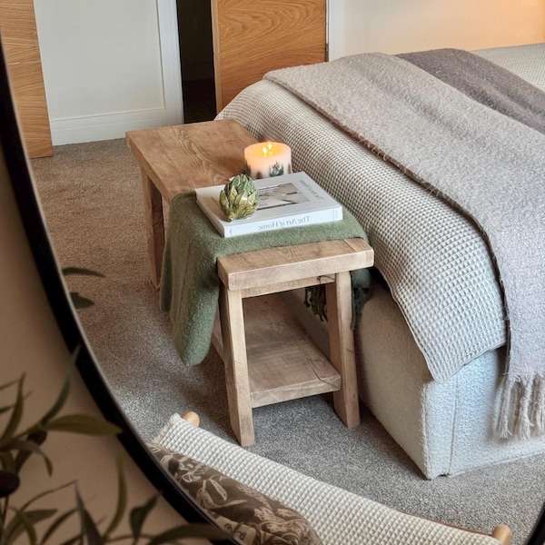 以下が含まれることがあります： 棚付きの木製のサイドテーブル、緑色の毛布、タイトルが「The Art of Home」の本、そしてその上に置かれたキャンドル。テーブルは、グレーと白の毛布をかけたベッドの隣にある寝室にあります。