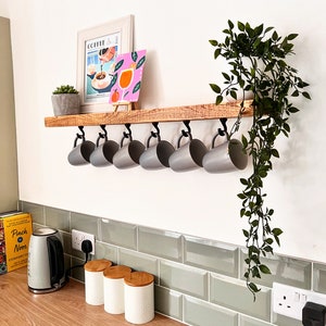 May include: A rustic wooden shelf with metal hooks holding grey mugs. The shelf is mounted on a white wall with a green plant hanging down. There are three white ceramic canisters with wooden lids on the counter below the shelf.