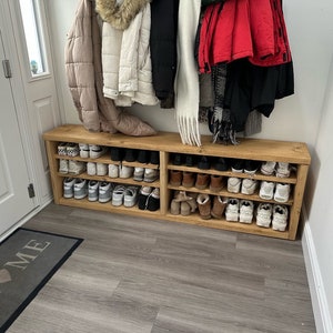May include: A rectangular wooden shoe rack with two horizontal shelves, featuring a natural light brown wood grain finish. The unit is filled with various pairs of trainers, boots, and casual footwear arranged neatly across the open shelving tiers.