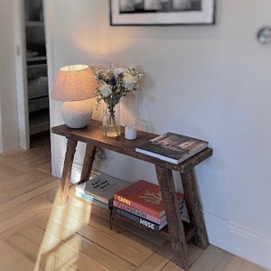 May include: A wooden bench with a white lamp, a vase of white flowers, and a candle on top. There are several books stacked on the lower shelf, including "Eat, Drink, Nap", "Capri", "Palm Beach", and "Mykonos Muse".