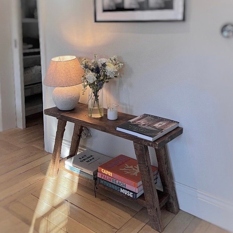 May include: A wooden bench with a white lamp, a vase of white flowers, and a candle on top. There are several books stacked on the lower shelf, including "Eat, Drink, Nap", "Capri", "Palm Beach", and "Mykonos Muse".