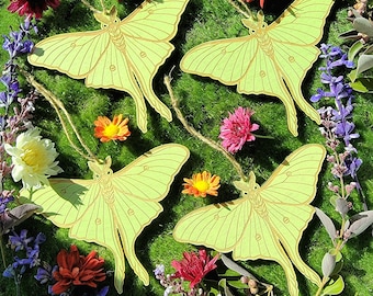 May include: Four green and yellow paper cutouts of luna moths with brown veins, hanging from twine against a background of green grass and colorful flowers.