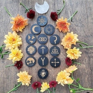 Op de afbeelding: Een set van 15 ronde zwarte houten tokens met gouden symbolen en ontwerpen. De tokens zijn in een cirkel op een houten oppervlak gerangschikt. De symbolen omvatten een halve maan, een ster, een zon, een boom, een vogel, een cirkel met drie stippen, een vraagteken, een knoop, een pijl, een letter Y, een kruis en een oog.