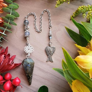 May include: A silver and grey crystal pendulum with a silver chain, a silver angel wing charm, and a silver mandala charm. The pendulum is on a wooden surface with flowers.