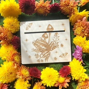 Könnte beinhalten: Eine Holzplakette mit einem lasergravierten Design eines Frosches, der auf einem Pilz sitzt, mit einem Schmetterling und Sternen. Die Plakette ist von gelben, roten und orangefarbenen Blumen umgeben.