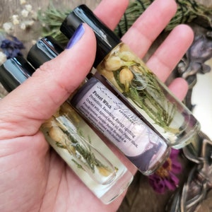 Pu&ograve; includere: Quattro bottiglie di vetro riempite con un liquido trasparente e prodotti botanici. Le bottiglie sono etichettate con "Forest Witch Apothecary" e elencano gli ingredienti: olio di nocciolo di albicocca, pepe nero, incenso, bacca di ginepro, bergamotto e gelsomino. Le bottiglie sono tenute in una mano.