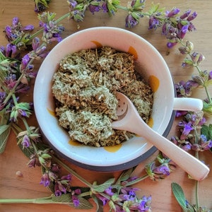 May include: A white ceramic bowl filled with a green herbal blend. The bowl has a yellow and white pattern and is sitting on a wooden surface. A wooden spoon with a star design is resting in the bowl. The bowl is surrounded by purple sage flowers.