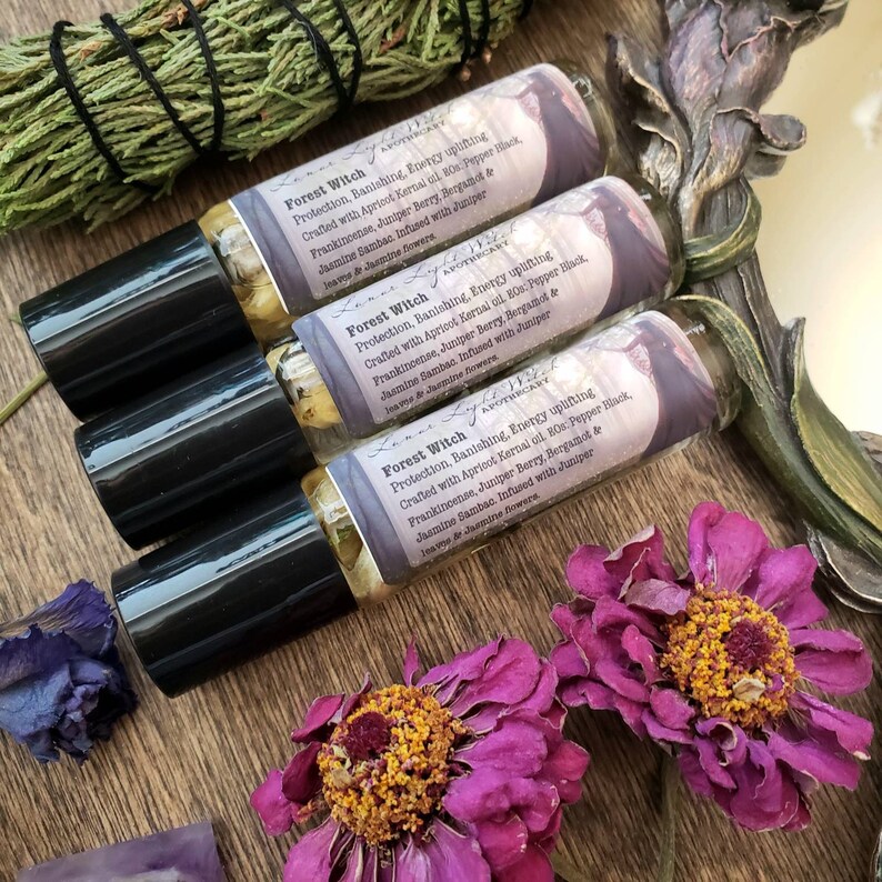 Pu&ograve; includere: Tre bottiglie di vetro con tappi a rullo neri e etichette che recitano "Forest Witch Protection, Banishing, Energy uplifting Crafted with Apricot Kernal oil, BOs: Pepper Black, Frankincense, Juniper Berry, Bergamot & Jasmine Sambac. Infused with Juniper leaves & Jasmine flowers" Le bottiglie sono su una superficie di legno con fiori secchi e un pezzo di ametista.