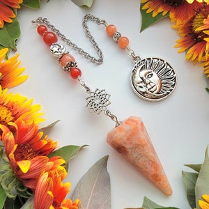 May include: A sun and moon pendant dangles from a silver chain with orange and red beads. The pendant is attached to a pointed orange crystal pendulum.