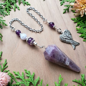 May include: A purple amethyst pendulum with a silver chain and angel wing charm. The pendulum features a faceted crystal point and is adorned with amethyst and white beads. The background is a wooden surface with greenery.