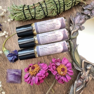 Pu&ograve; includere: Tre bottiglie di vetro con tappi neri ed etichette che recitano "Forest Witch Apothecary" e elencano ingredienti come bacche di ginepro, incenso, sandalo e pepe nero. Le bottiglie sono impilate su una superficie di legno con fiori viola e un cristallo viola.