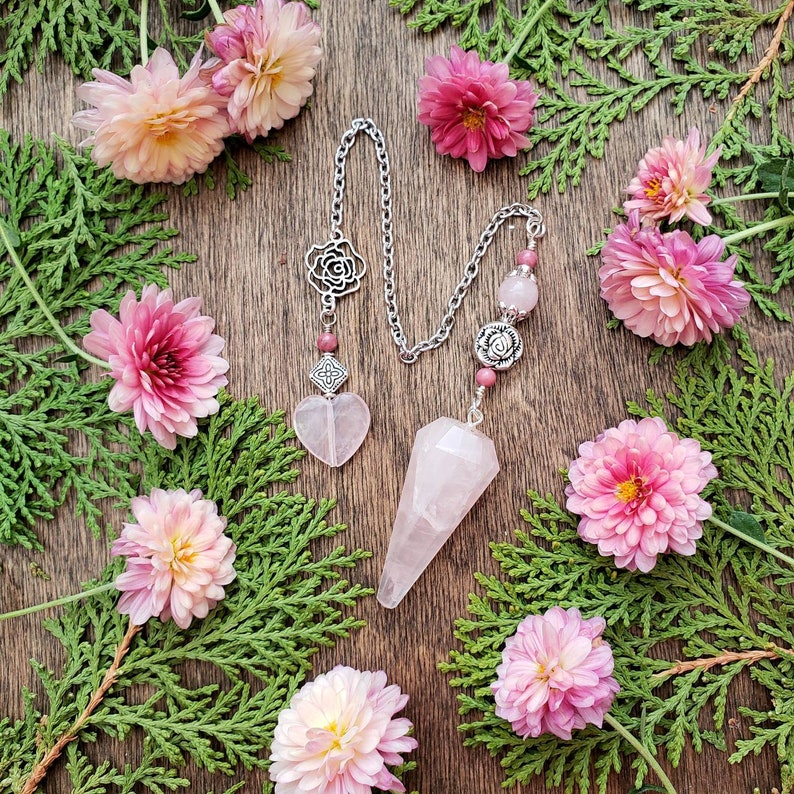 May include: A pink crystal pendulum with a silver chain and rose-shaped charms. The pendulum is surrounded by pink flowers and green foliage.