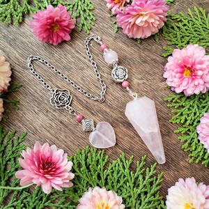 May include: A pink rose quartz pendulum with a silver chain and rose-shaped charms. The pendulum is on a wooden surface with pink flowers.
