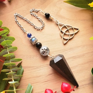 Puede incluir: Péndulo de obsidiana negra con una cadena y cuentas plateadas, incluyendo una piedra azul, una piedra negra y un dije plateado con un diseño de árbol. La cadena está unida a un dije de nudo celta plateado.