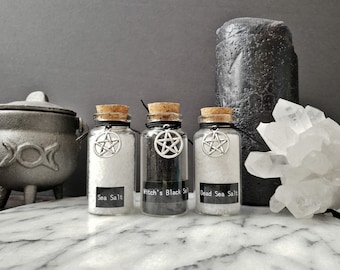Puede incluir: Tres pequeñas botellas de vidrio con tapas de corcho y una cuerda negra atada alrededor del cuello. Cada botella contiene un tipo diferente de sal: sal marina, sal negra de bruja y sal del Mar Muerto. Cada botella tiene un amuleto de pentagrama plateado colgando de la cuerda.