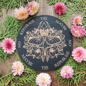 Könnte beinhalten: Ein schwarzes Ouija-Brett aus Holz mit goldenen Buchstaben und einem Motten-Design. Das Brett hat die Wörter "Ja", "Nein", "Vielleicht" und "Umformulieren" am Rand.