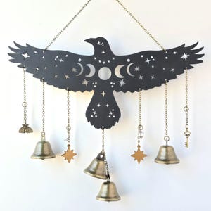 May include: Black metal raven wind chime with moon phases and stars cutouts. The chime has six hanging chains with bells, stars, a key, and a moth charm.