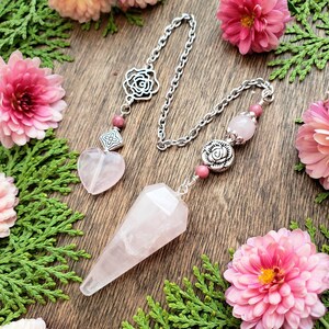 May include: A pink rose quartz pendulum with a silver chain and rose-shaped charms. The pendulum is hanging from a wooden surface with pink flowers.