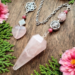 A rose quartz pendulum with a silver chain and rose-shaped charms. The pendulum is a pointed crystal with a heart-shaped rose quartz charm attached to the chain.